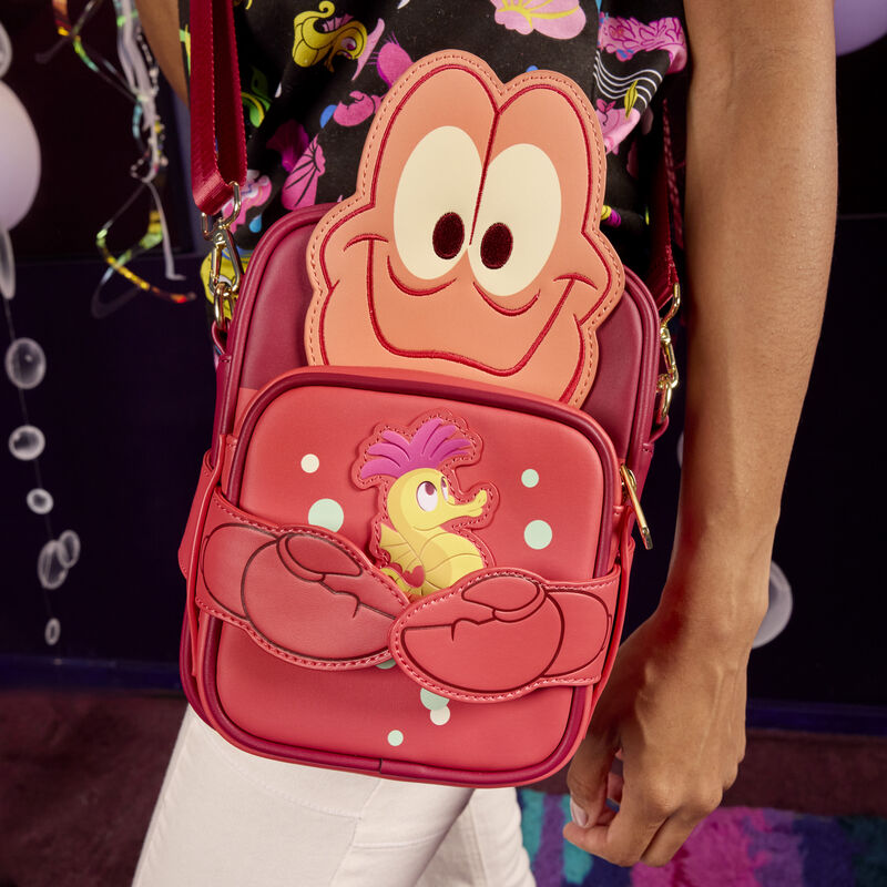 Person facing to the side, wearing the Loungefly Little Mermaid Sebastian Crossbuddies bag, featuring Sebastian as an appliqué on the red crossbody and holding a coin pouch with a seahorse on it between his claws. 